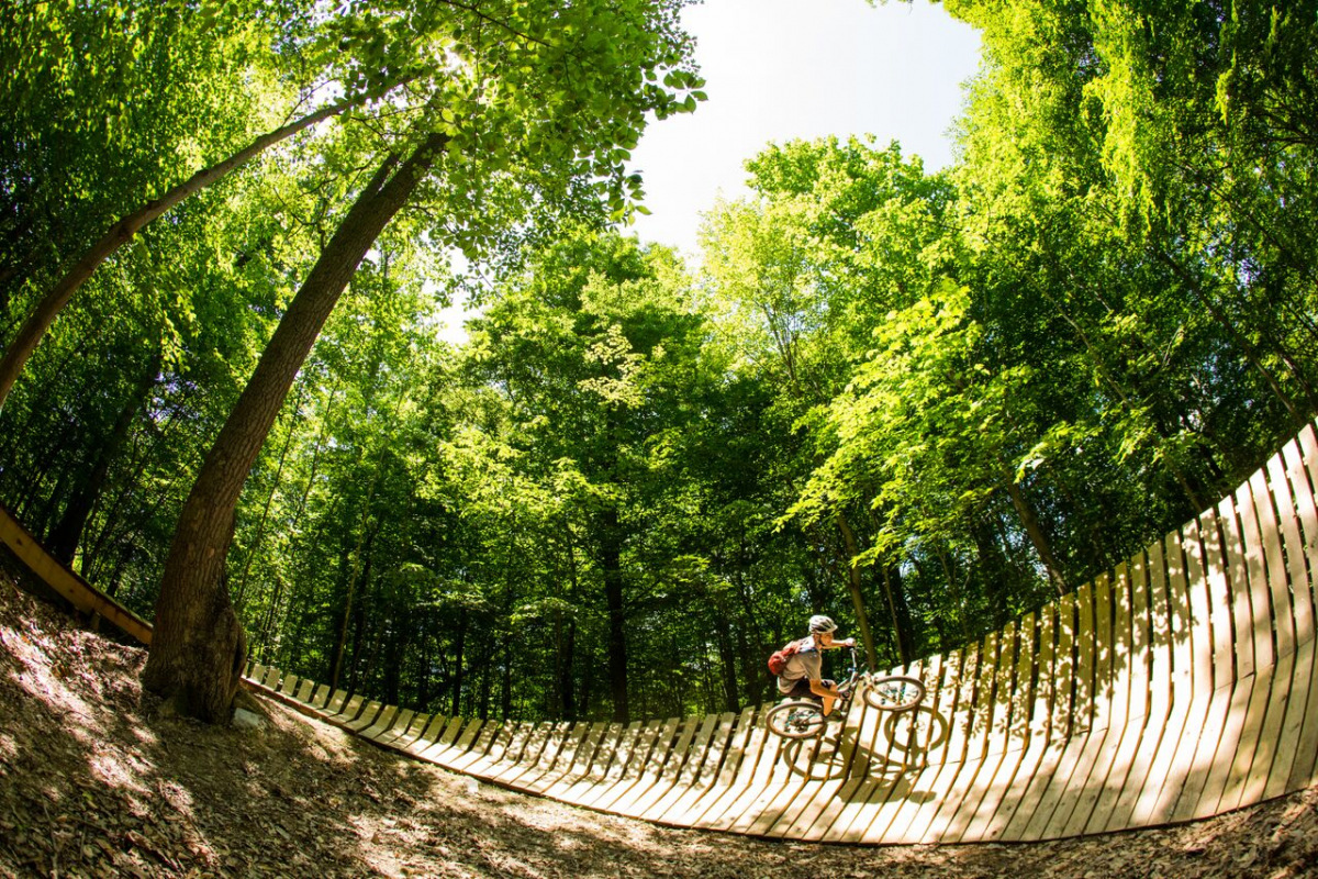 A person on a mountain bike rides along a curved wooden ramp next to some tall trees.
