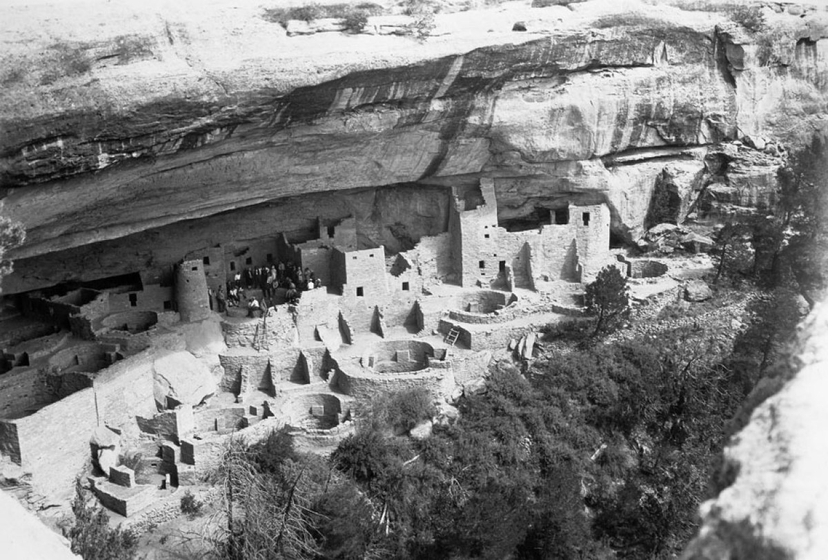 An aerial view of ancient buildings carved into the side of a desert canyon and a cluster of tourists sitting among the ruins with bushes and trees further into the ravine.