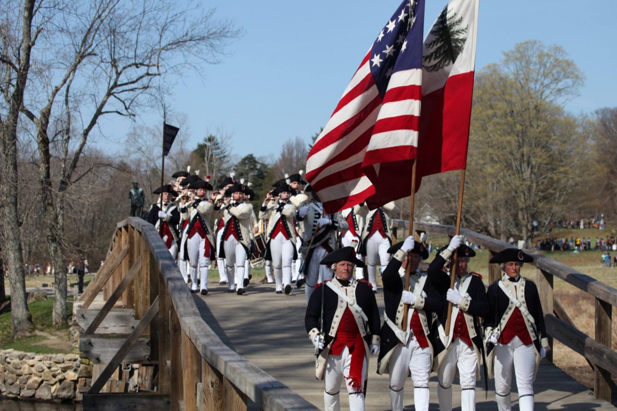 On a crisp autumn day, a large group of living historians in era-appropriate uniforms march across a wooden bridge with flutes, drums, replica weaponry and large flags while observers watch from green fields in the background.