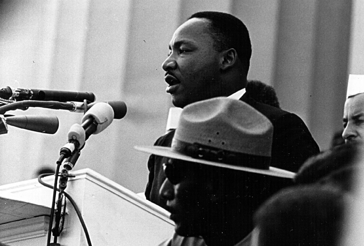 Historic black and white photo of Dr. King speaking into several microphones on a podium with a white columned building behind him.