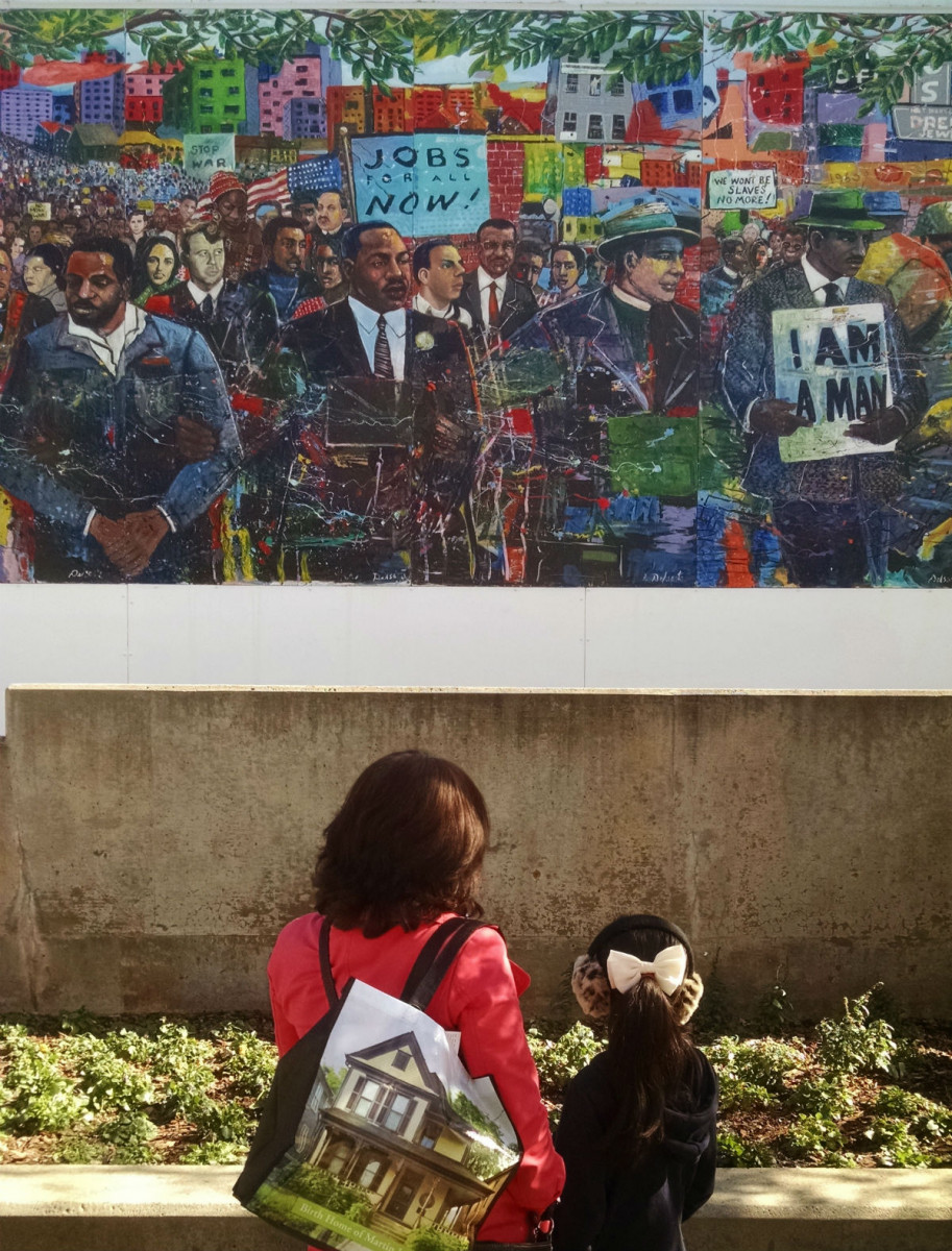 An African American woman and little girl stand in front of a wall painted with a mural showing civil rights leaders marching.