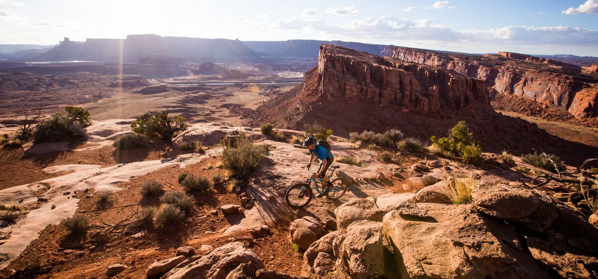 A white man wearing a helmet rides a bike across a red rock plateau with a rugged desert landscape in the background.