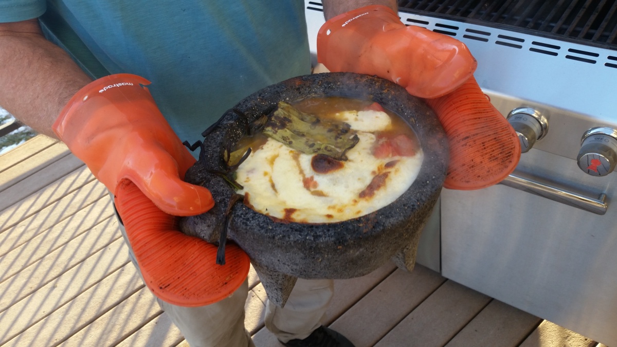 A man in a blue shirt holds a stone bowl in front of him. It is covered in melted cheese and he wears orange oven mitts.