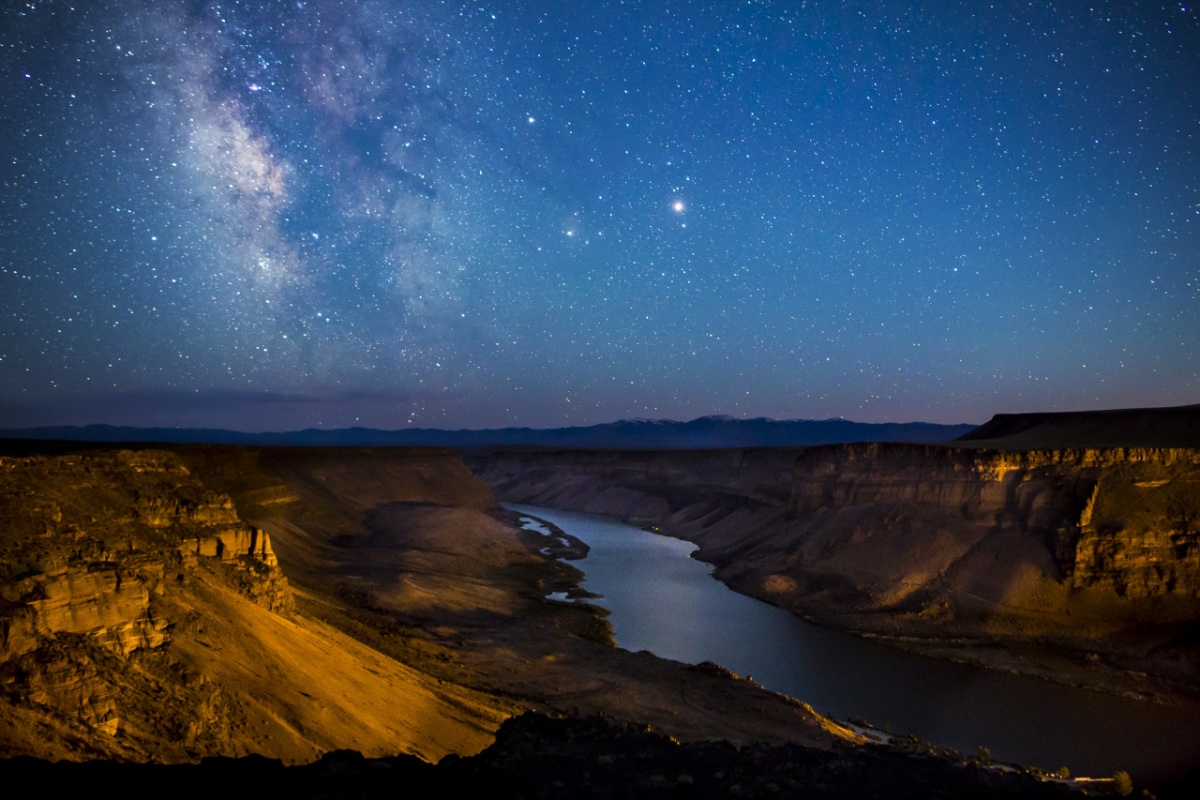 Bright orange rock and deep blue river glow in the light of a bright blue and purple star-filled sky.