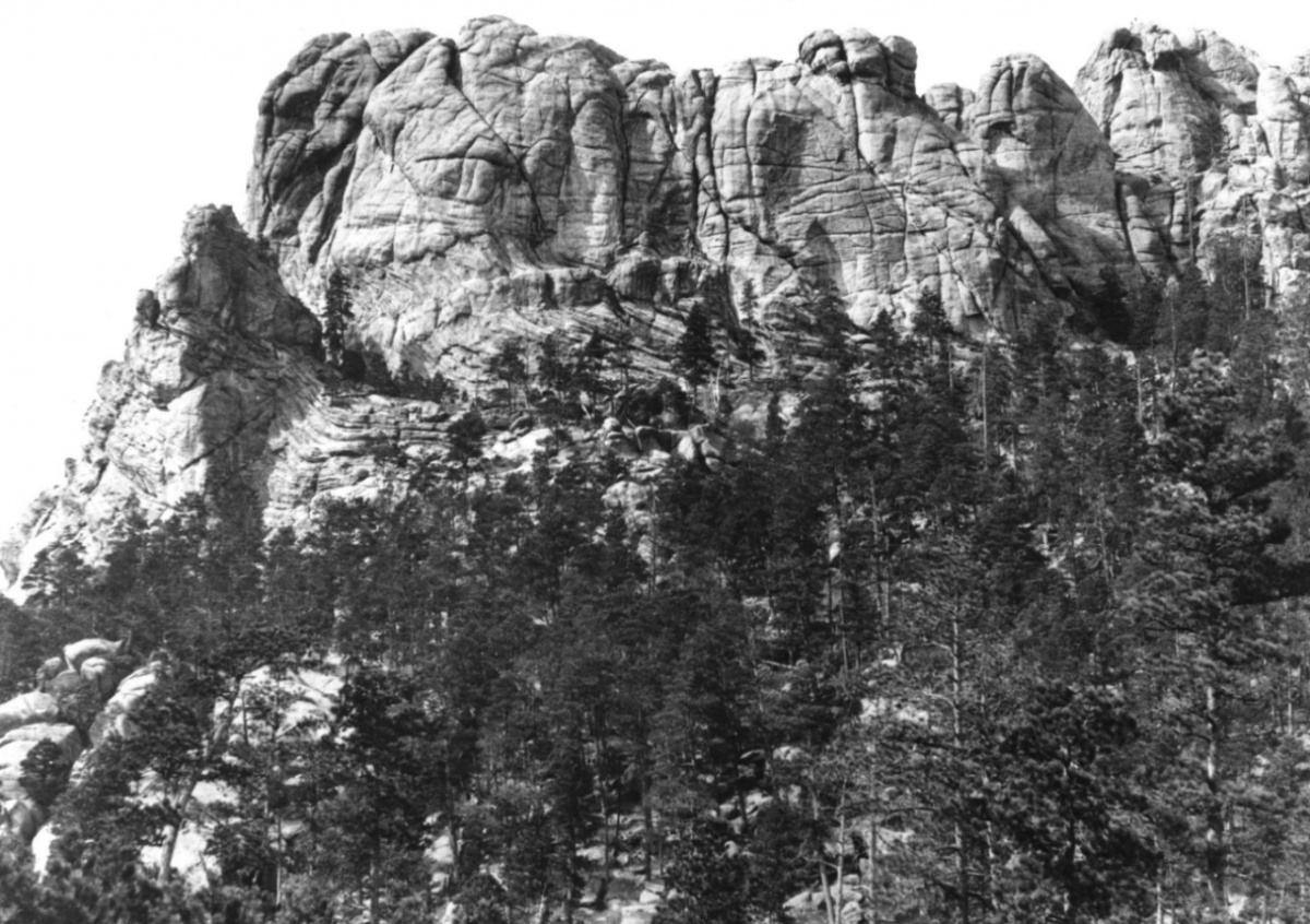 Photo of Mount Rushmore before the faces were carved in it. A tall rocky cliff with trees at the base.