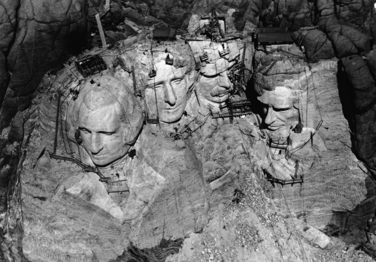 A historic aerial photo of Mount Rushmore under construction. The four heads of the memorial are covered in cranes and workers.