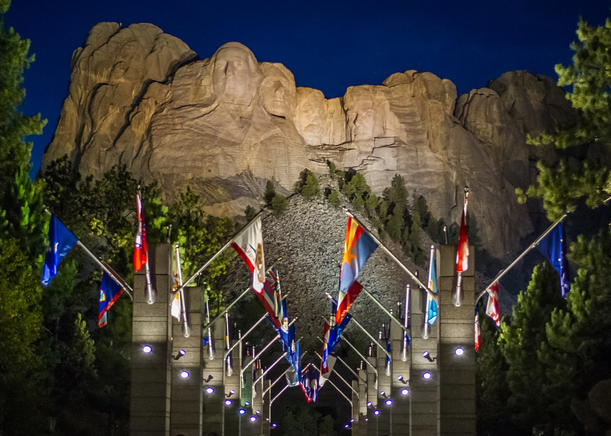 An outdoor walkway lined with state flags moves towards the sculpture on Mount Rushmore.