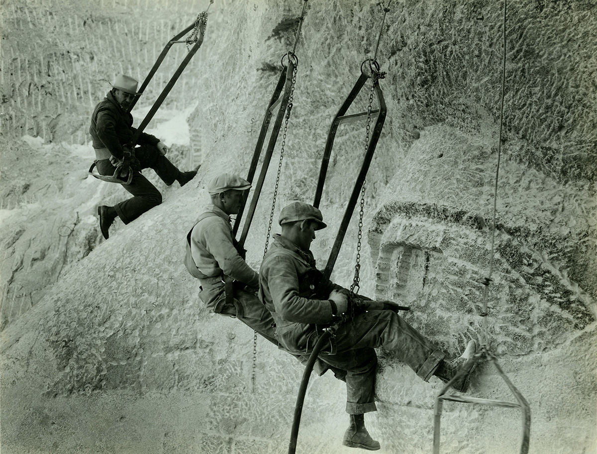 A historic photo of workmen hanging by ropes on the side of Mount Rushmore using tools to carve the eyes of a face into the stone.