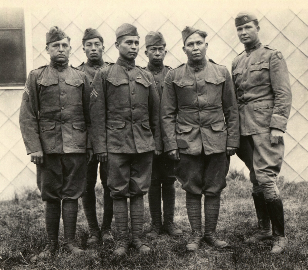 A historic photo of a small group of native american men in world war one uniforms posing in front of a wooden building.