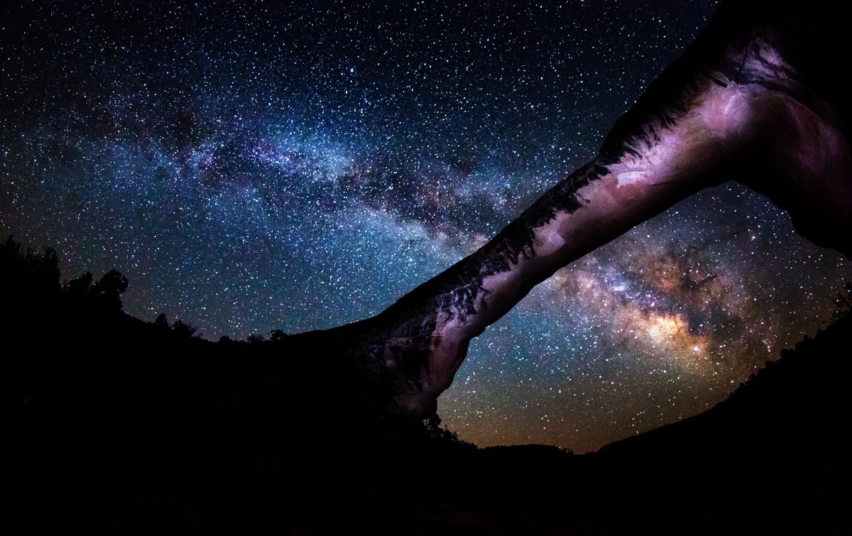 The Milky Way and a dark starry night illuminate a rock bridge. Photo taken from below bridge looking up into sky.