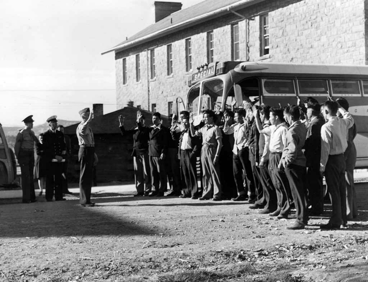 A black and white historic photo of a group of 29 native american men stand by a bus in front of a large stone building taking an oath from a military officer.
