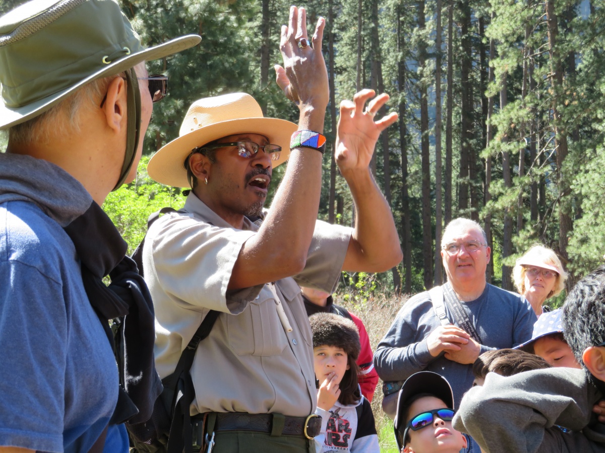 Shelton Johnson, an African American man in a park ranger uniform and hat, talks to a small group of people with a forest in the background.