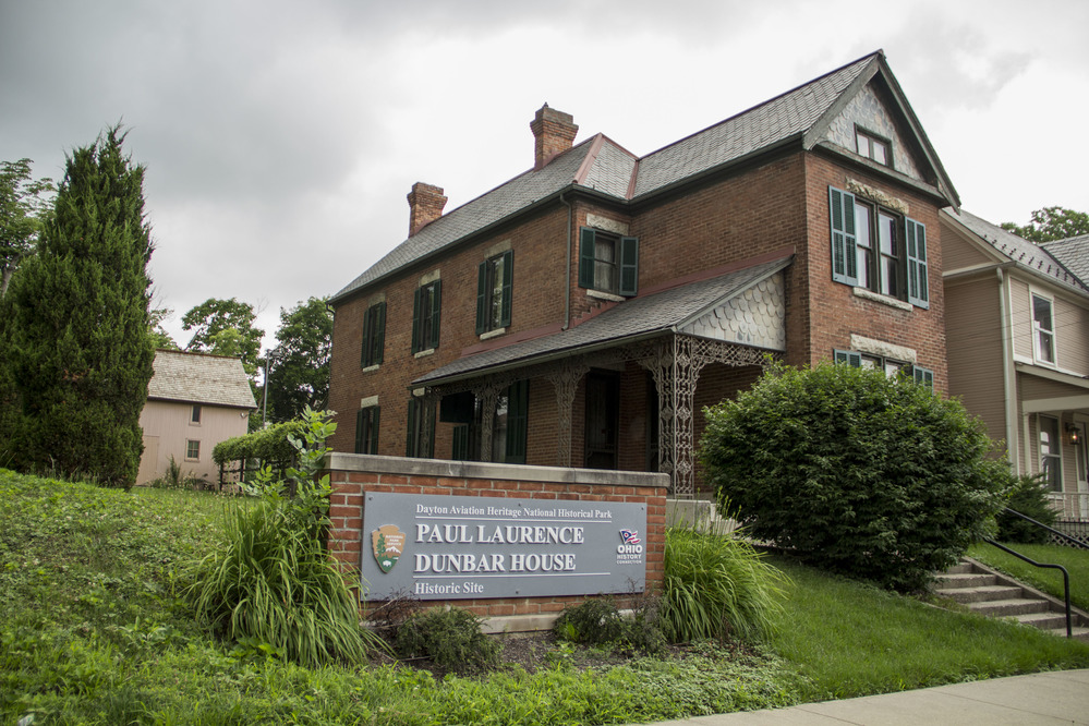 Against a backdrop of gray clouds, a federalist-style brick building is framed by green foliage and a large National Park Service sign.