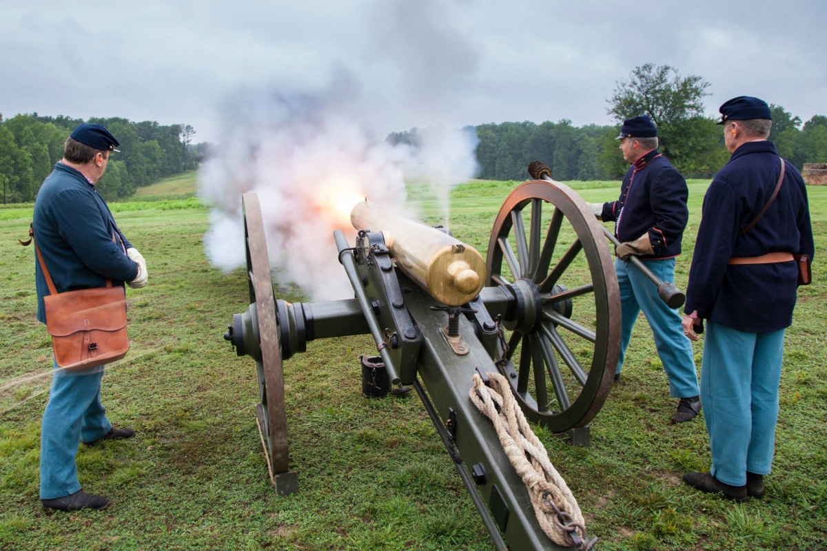Three men dressed in uniforms fire a large gold cannon. Large white smoke clouds follow the shot.