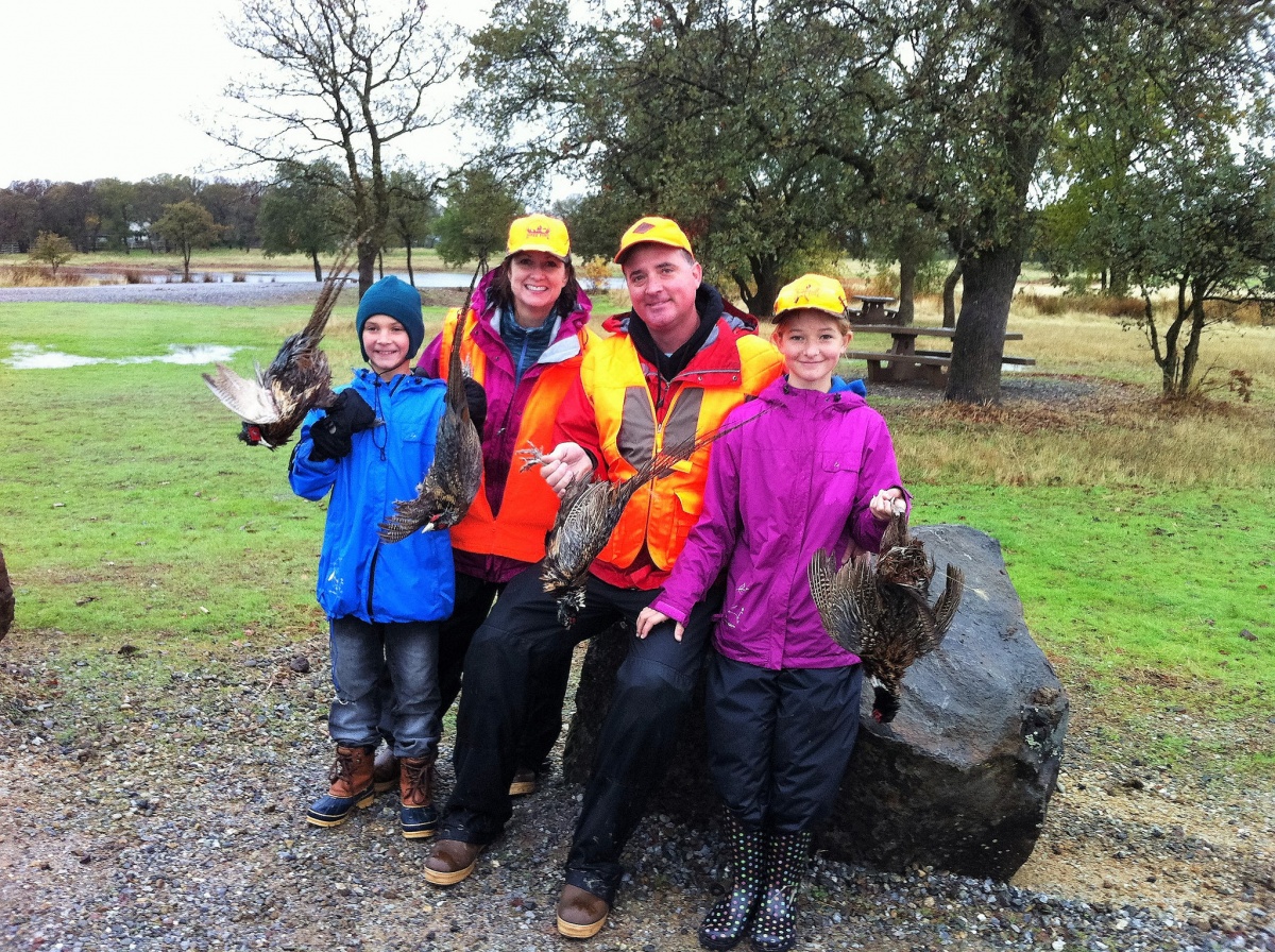 A mother, father, son and daughter smile for a photo and hold up pheasants they have just hunted. They are all wearing bright colored coats and hats.