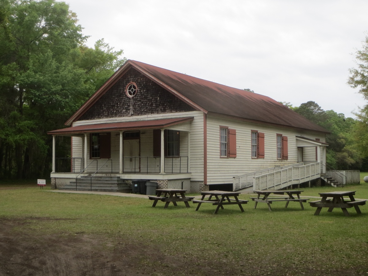 An old, long wooden building with a porch stands in a yard surrounded by trees.