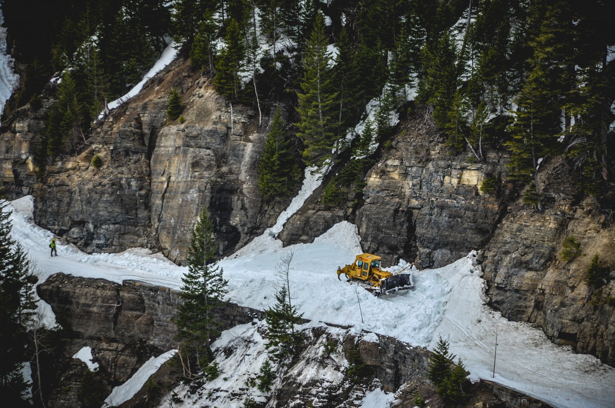 A yellow bulldozer plow snow on a narrow road built on a ledge of a mountain.
