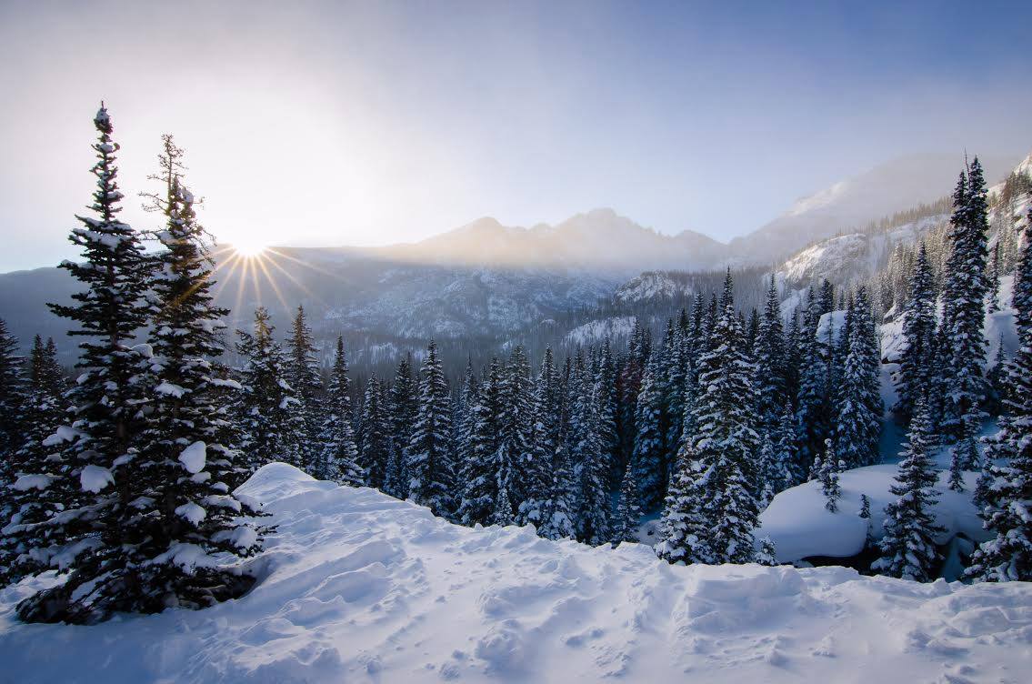 The sun peeks over the mountain, revealing snow-covered landscape of trees and mountains