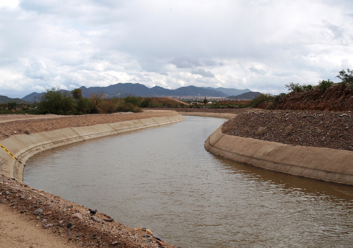 A canal filled with water curves through a desert landscape with a city and mountains on the distant horizon.