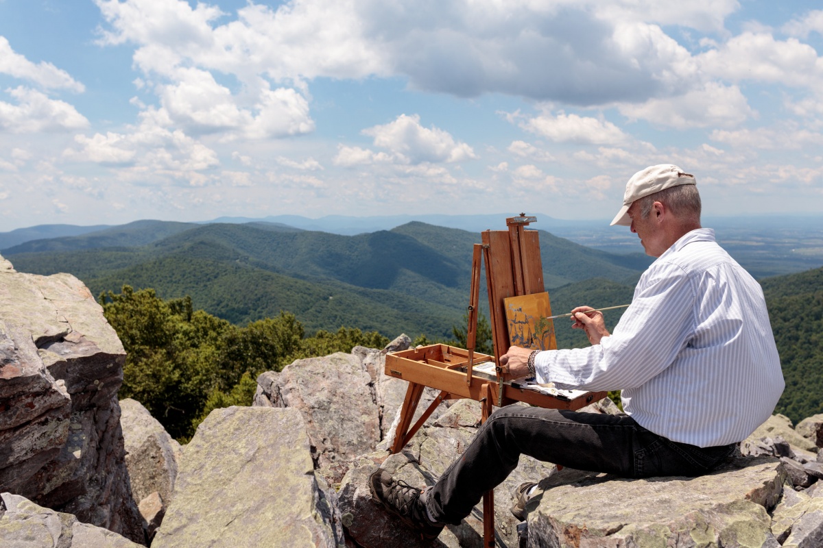 An artist sits on rocks with his easel painting the rolling green hills in front of him.
