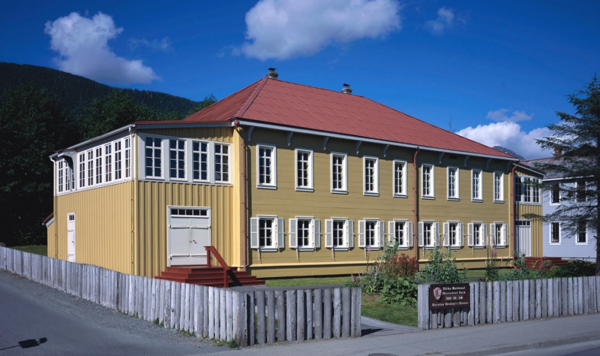 A wide, tan two story building with a red roof stands in a yard bordered with a wooden fence.