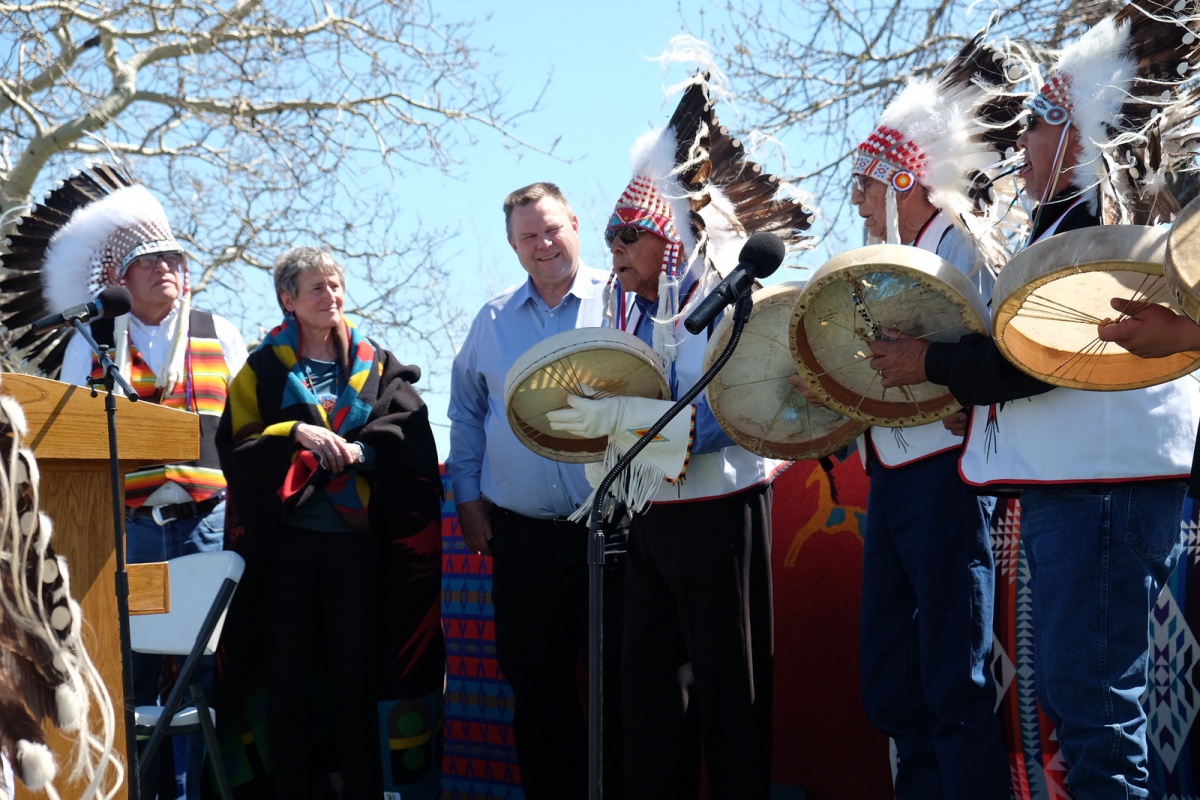 Secretary Jewell stands on a stage as Native Americans in traditional clothes play drums.