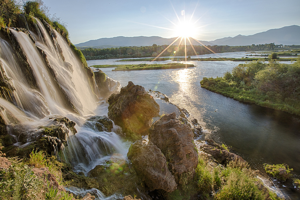 A waterfall runs over a small cliff and flows down to a wide river running past a forest with mountains in the distance under a bright sunny sky.