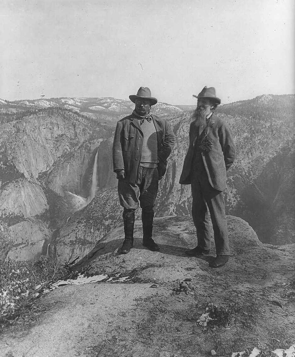 Theodore Roosevelt and John Muir stand on a rock cliff overlooking a valley and waterfall.