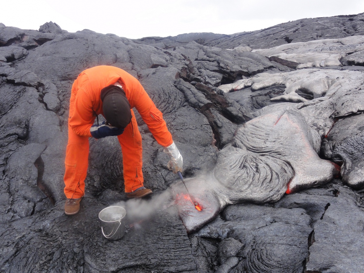 A scientist in orange protective gear dips a tool into red hot lava, extracting it from surrounding grey rock from a volcano.