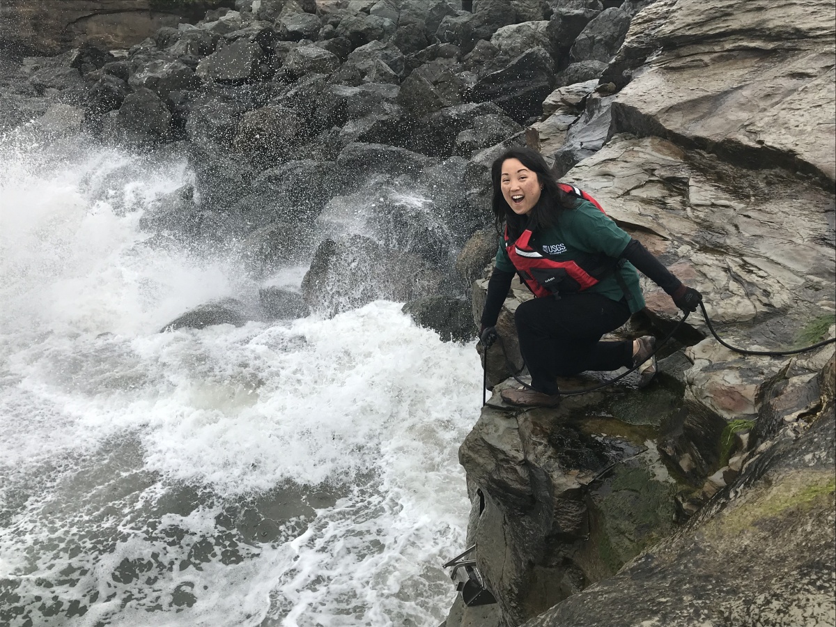 A smiling Asian American woman wearing safety gear kneels on a rocky shoreline lowering a scientific instrument into the ocean waves below her. 