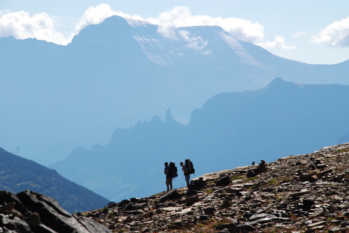 Two people with large backpacks stand in the center of a rocky mountain, with mountains stretching into the blue sky in the distance behind them.