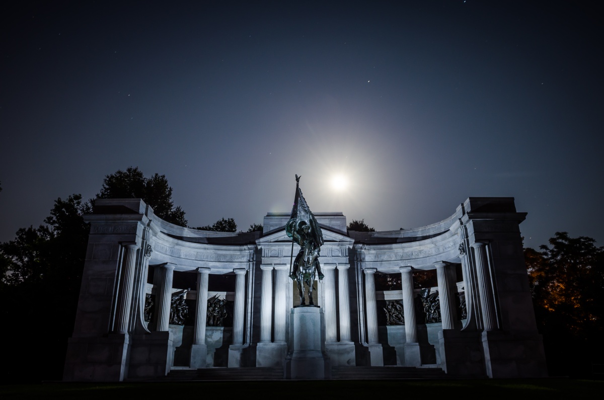 The moon shines bright in a dark and starry sky over a large white monument. 