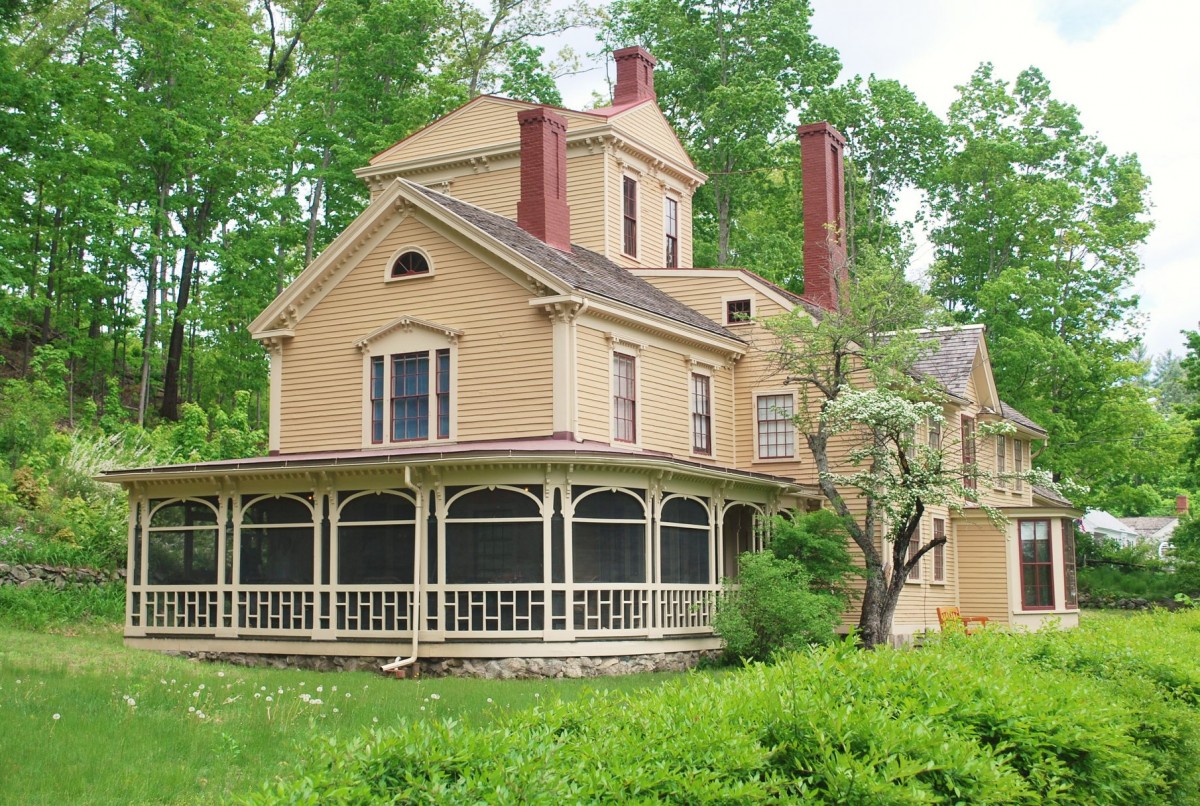 A yellow, wood-panelled two-story house with three tall red brick chimneys sits amongst bright green leafs and shrubbery.