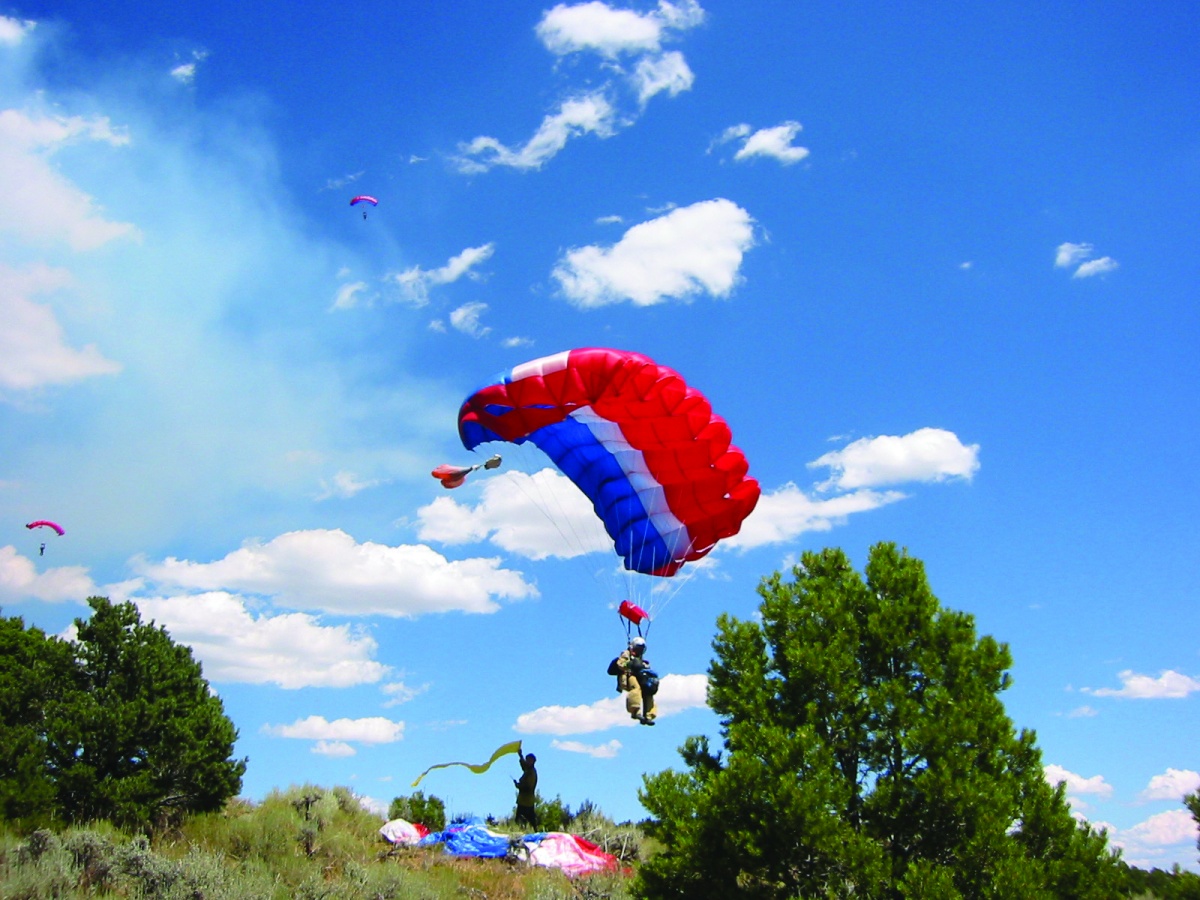 Two firefighters wearing protective gear hang by parachutes in the sky as they come in for a landing on a grassy hill.