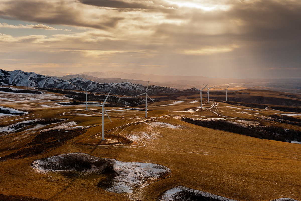 Sun rays shine through clouds onto a light, bright brown field filled with wind turbines. Snow covered patches and peaks line the area.