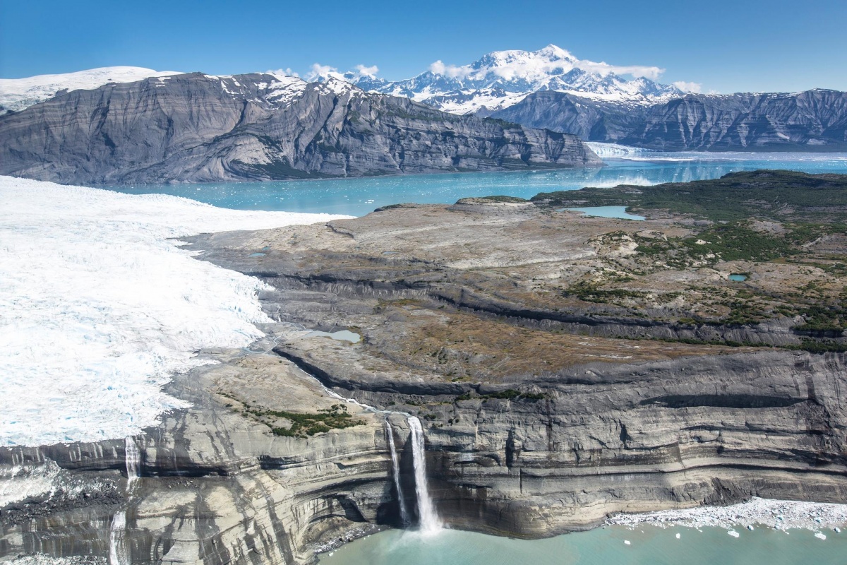 An aerial photo of mountain ranges and massive glaciers cover a wide landscape running down to blue inlets.