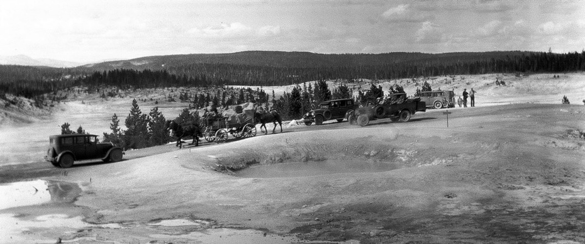 An old black and white photo of early automobiles and people near several bubbling hot springs.