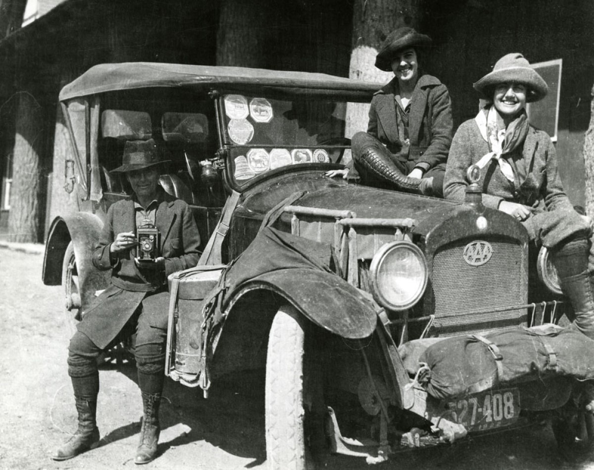Three women with wide-brimmed hats and long coats sit smiling on an historic automobile adorned with stickers and covered in dust.
