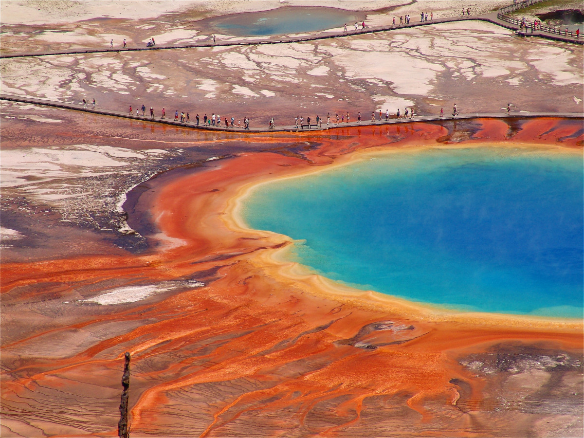 A view looking down at people walking along a long wooden boardwalk past a large blue pool of water bordered by bright colorful lines of dirt.