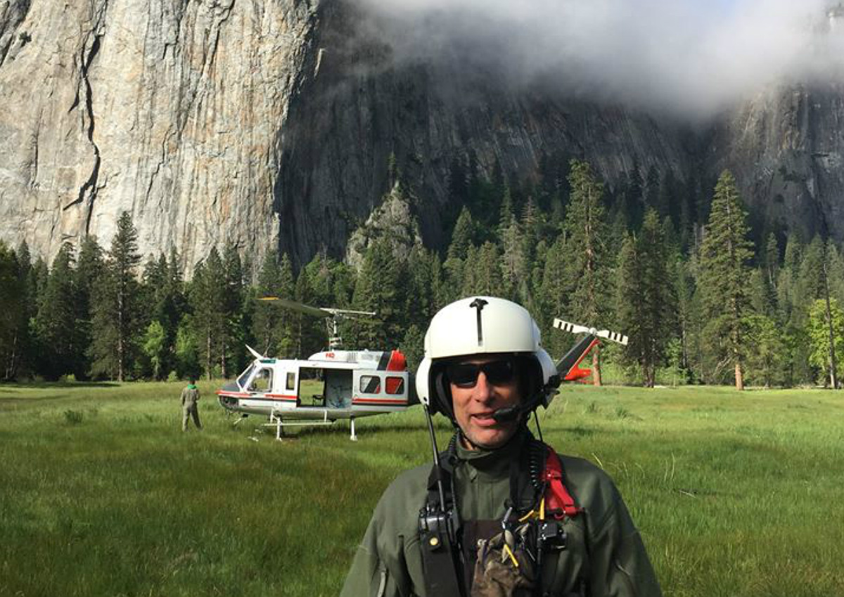 A white man in a helmet and flight suit stand near a helicopter in a grassy meadow with a granite cliff behind him.