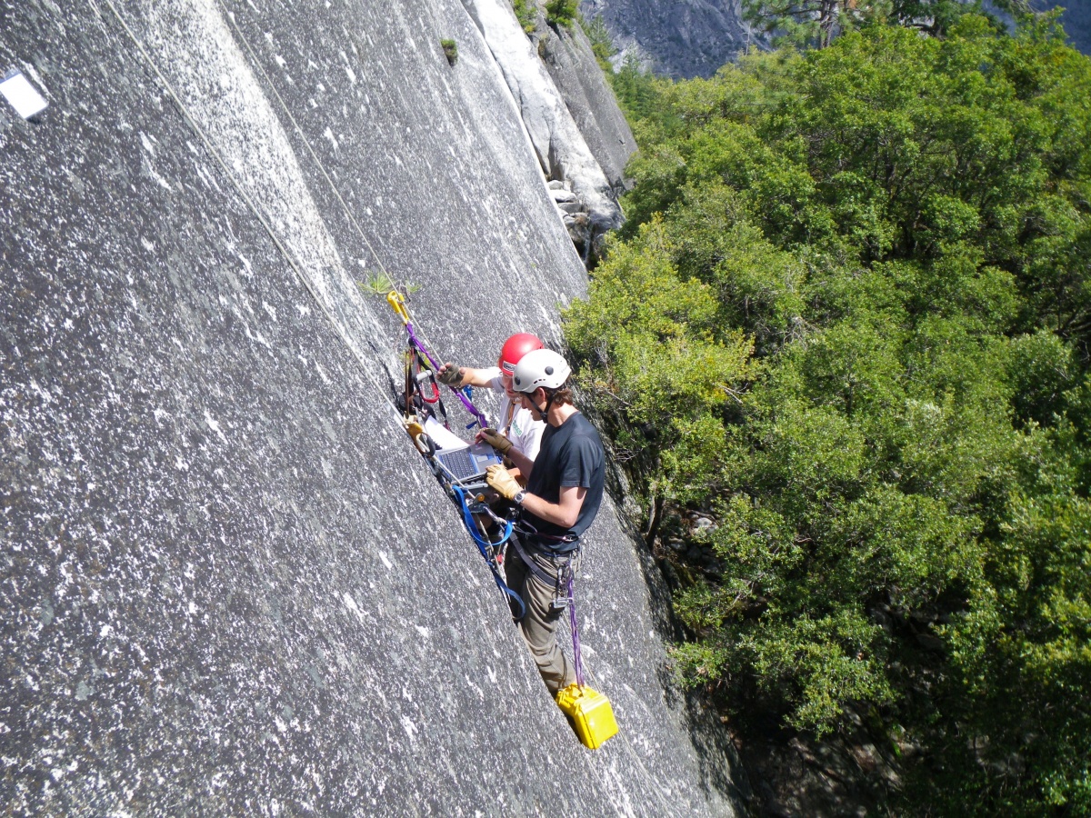 Two people wearing climbing gear hang from ropes against a rock wall.