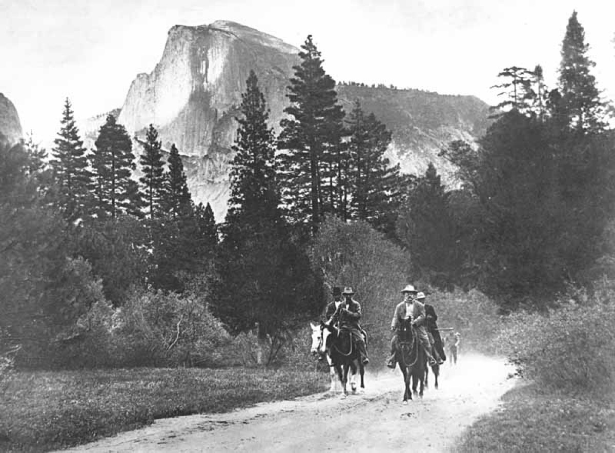 A group of men on horseback tread on a dusty trail lined with brush and trees in front of Half Dome in Yosemite National Park.
