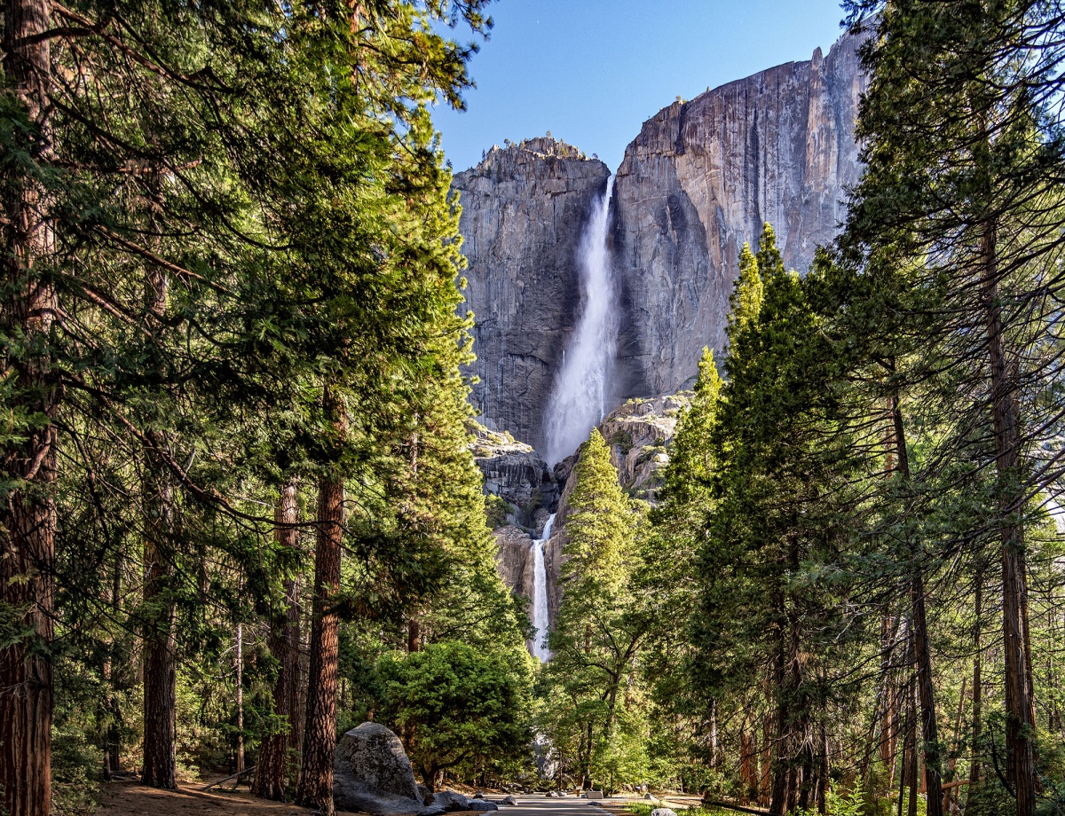 A tall waterfall flows over a high gray rock cliff with a forest at the base.