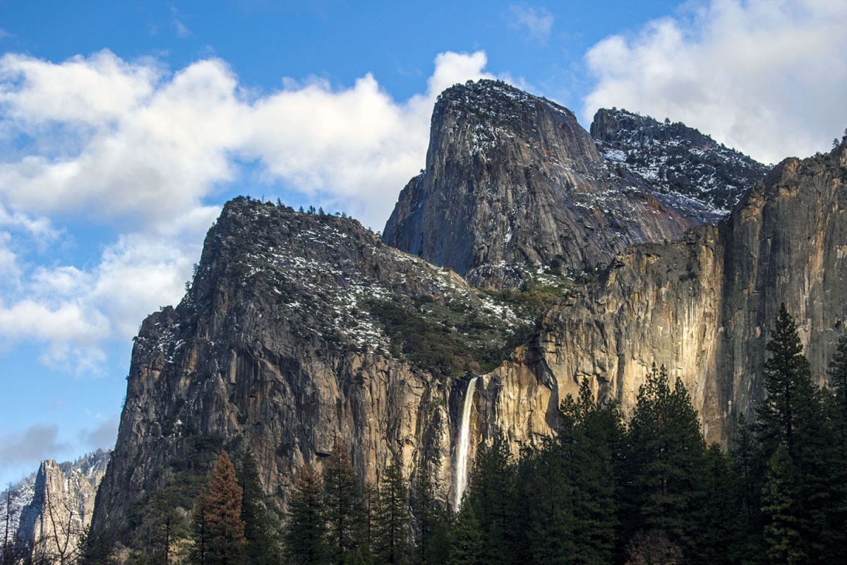 A waterfall tumbles down the rock face of wall of granite mountains dusted with snow.