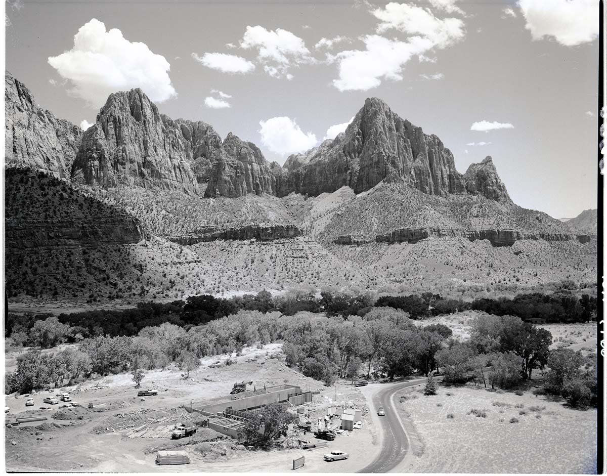 A twisting road and busy construction site with vintage cars and cement foundation sits at the foot of jagged desert formations spotted with brush under a cloudy but bright sky.