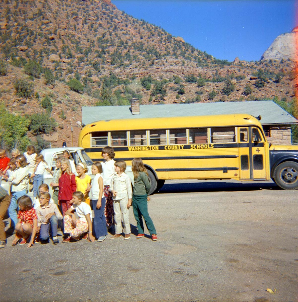 A group of young students gather together for a photo next to a vintage yellow school bus in front of desert hills dotted with brown dusty rocks and green shrubbery beneath a deep blue sky.