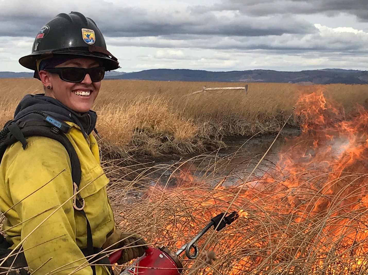 A firefighter wearing protective equipment, including a yellow jacket and brownish hard hat with the U.S. Fish and Wildlife Service logo, while smiling at the camera as fire burns brown, dry vegetation in the background.