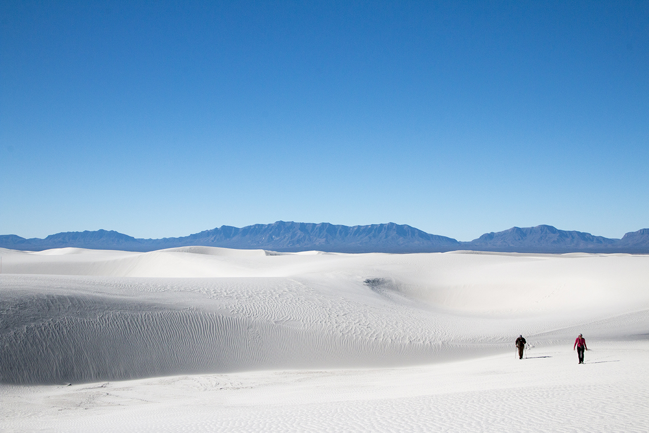 Two people walk along great wave-like dunes of gypsum sand.