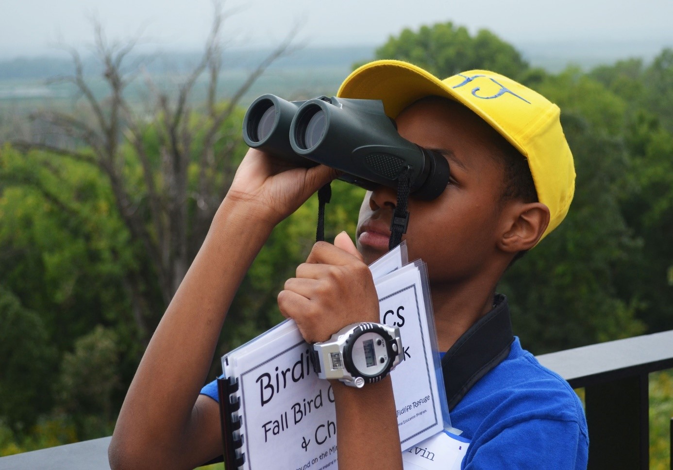 Child with binoculars, wearing a yellow hat and holding a book about birds.